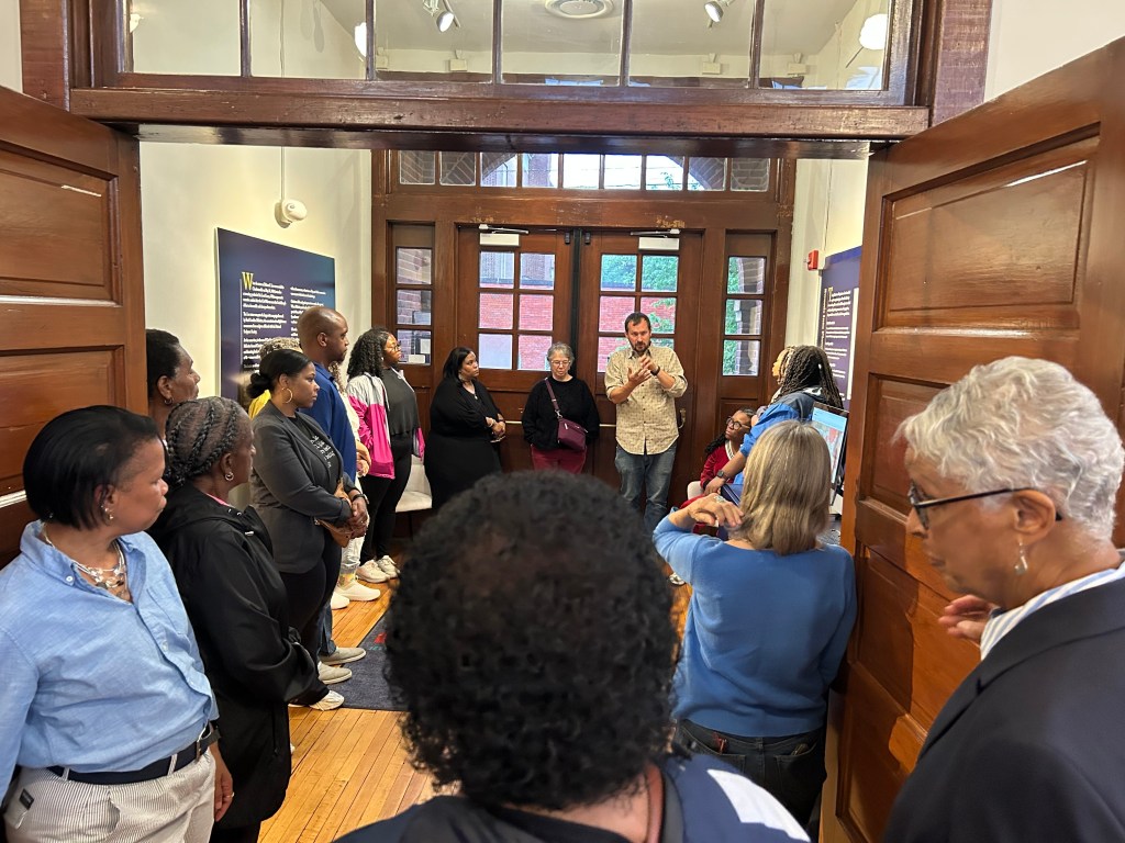 Several people are gathered in an entryway of a building, listening to a man who is standing in front of the entry doors speaking and gesturing with his hands.