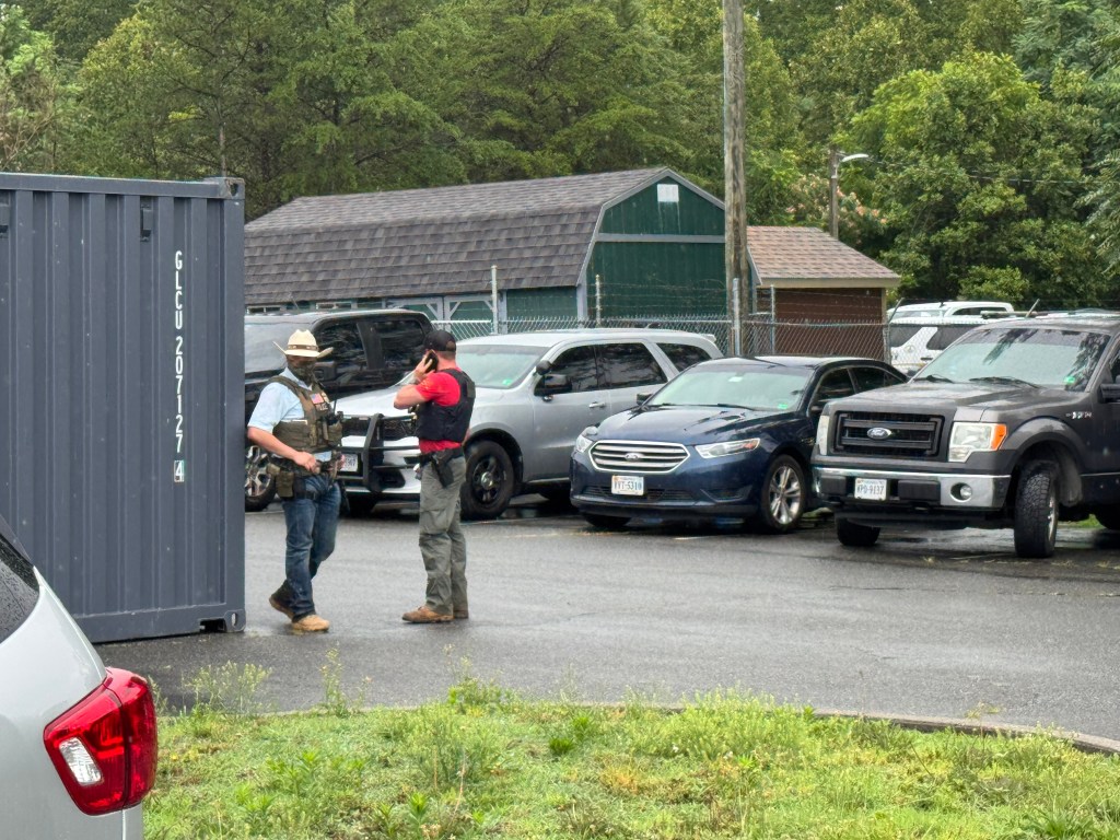 Two men in protective vests with gun holsters strapped to their belts stand by a dumpster in a parking lot. One wears a camouflage face mask and cowboy hat. The other is wearing a black baseball cap and speaking into a cellphone.
