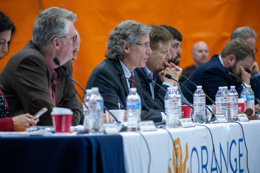 Several men in business suits sit at a table with microphones, water bottles and a white sign with the word "ORANGE" in blue lettering. The man at the center speaks into his microphone while the others listen.