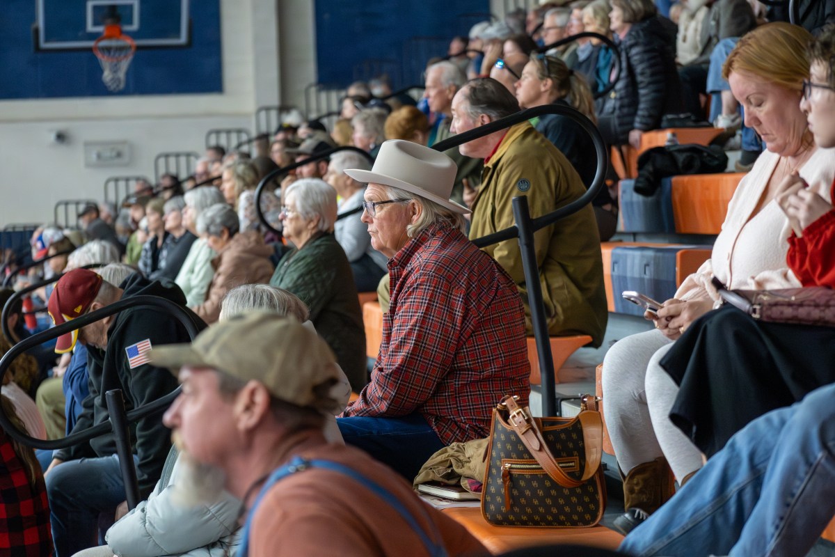 Many people fill a set of orange and dark blue bleachers in a high school gymnasium, including a central figure in a white, wide-brimmed hat and red plaid shirt. The audience members look straight ahead while listening to a speaker who is off-camera.