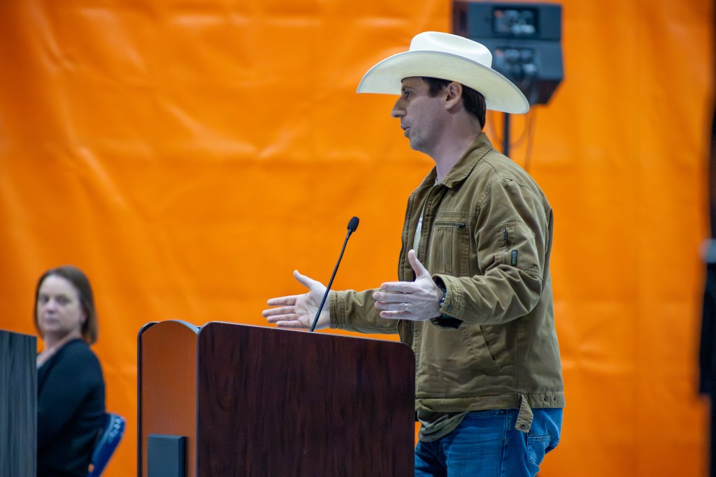 A man in a beige cowboy hat and light brown jacket speaks at a wooden podium against a bright orange background, with his arms partially outstretched.