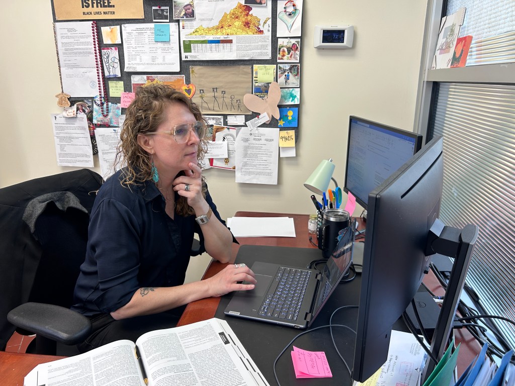 A woman in glasses sits at a desk with a large reference book to the side, a laptop and two monitors in front of her. A crowded bulletin board is behind her with fliers, pictures and documents, and a colorful map of Virginia tacked on.