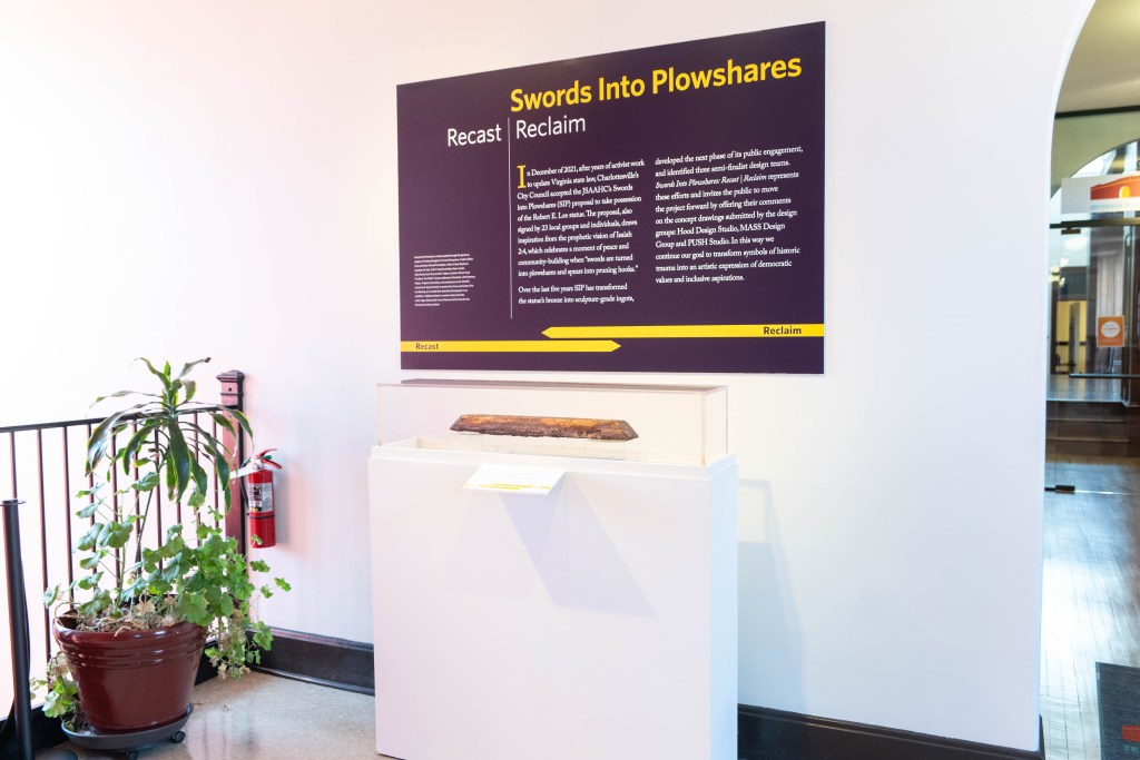 A piece of wood is displayed under glass at the entrance of an exhibit, with a large sign above it reading "Swords into Plowshares - Recast Reclaim"