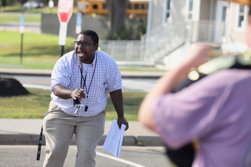 A man stands outside in a parking lot speaking and gesturing with his hands. High school students holding brass musical instruments stand facing him.