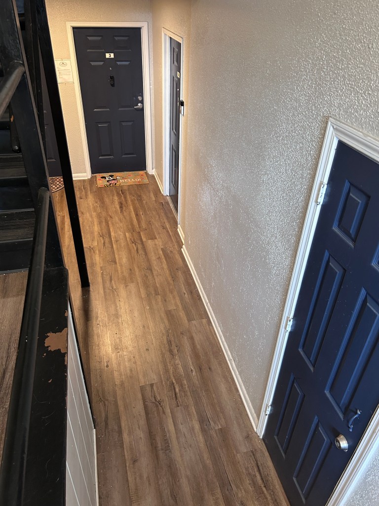 On the right, a hallway with clean, wood color floors, off-white walls and black doors, and hallway lights.