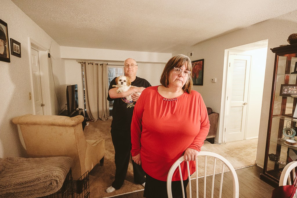 A woman stands at a chair with a small living room behind her, with a man holding a small dog in the background. Both are looking at the camera.