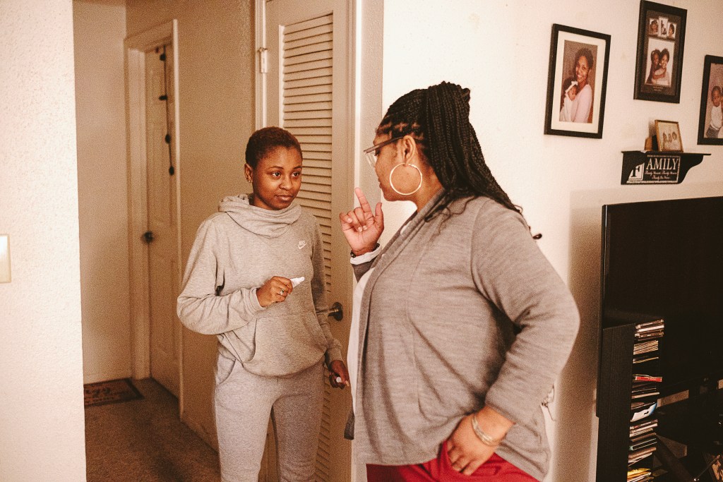 A person in a gray sweatsuit stands in the hallway of a home, with a woman in a gray sweater and large hoop earrings, facing away from the camera, speaking to her. To the side are family photos on the wall, a television and a rack of CDs or DVDs.