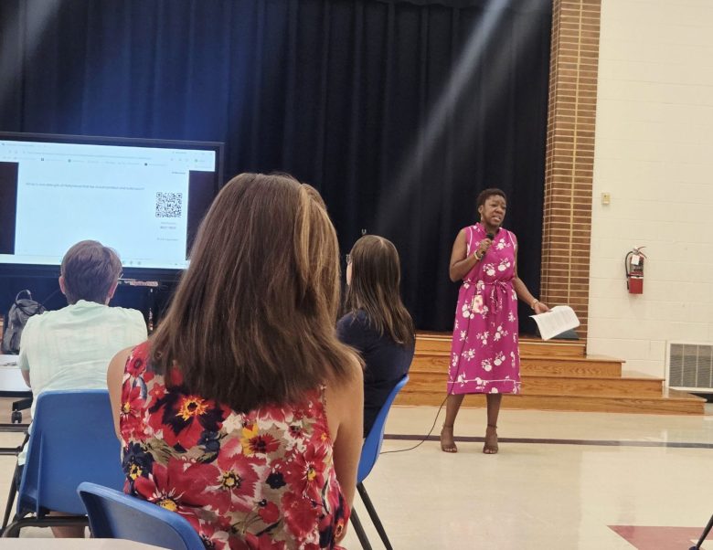 A woman is holding a microphone in her right hand and a few papers in her left, as she stands in front of several people in an auditorium.