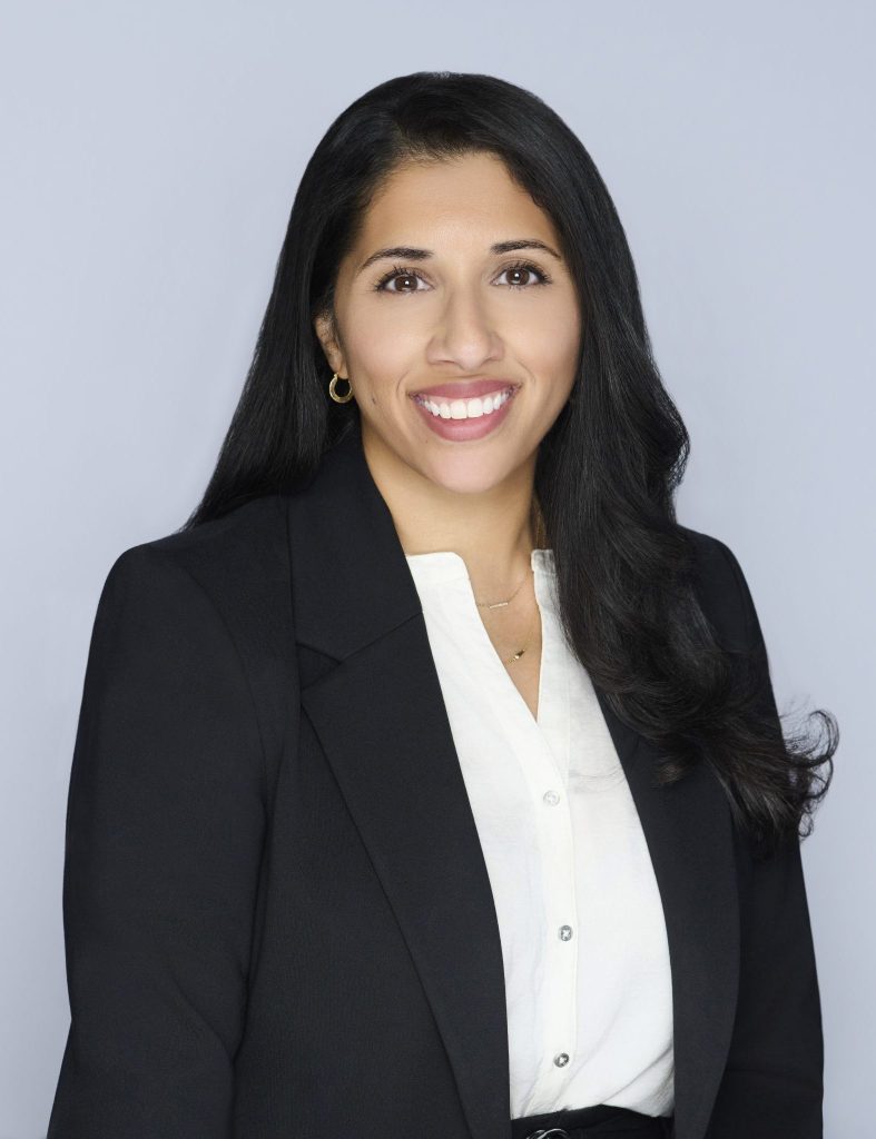 A woman in a black blazer and white button-down shirt smiles at the camera.