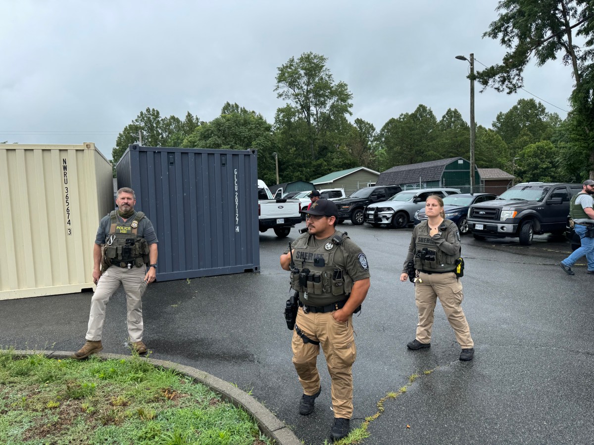 Three people in tactical great stand in a parking lot with cars and two temporary metal sheds behind them. There are two more people in the background. Two have vests that say "SHERIFF" and one has a vest that says "POLICE HSI"