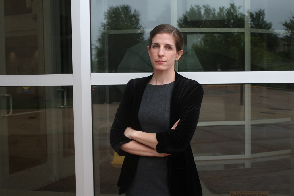 A woman in a black blazer and grey dress stands with her arms crossed in front of a building with mirrored windows.