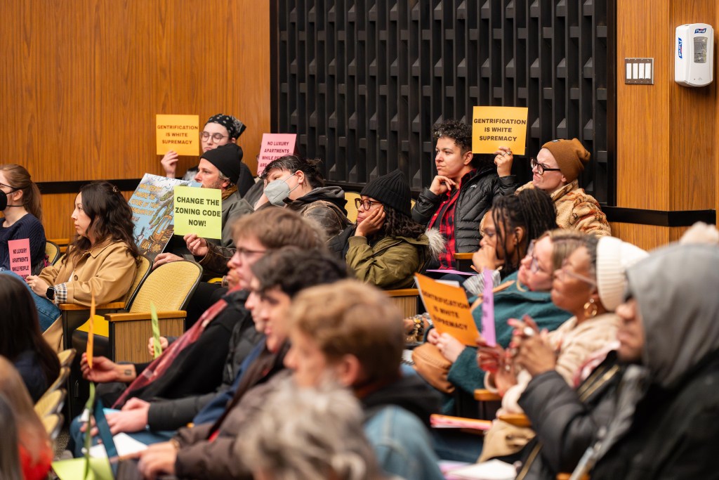 A close-frame photo of about 20 people in city council chambers, a mostly wood room with sound dampening foam on some of the walls. The group is multi-racial, and some of them hold up signs that say "gentrification is white supremacy," "change the zoning code now!," and "no luxury apartments while people are homeless" and "protect core Black neighborhoods." Most of them have unhappy expressions on their faces.