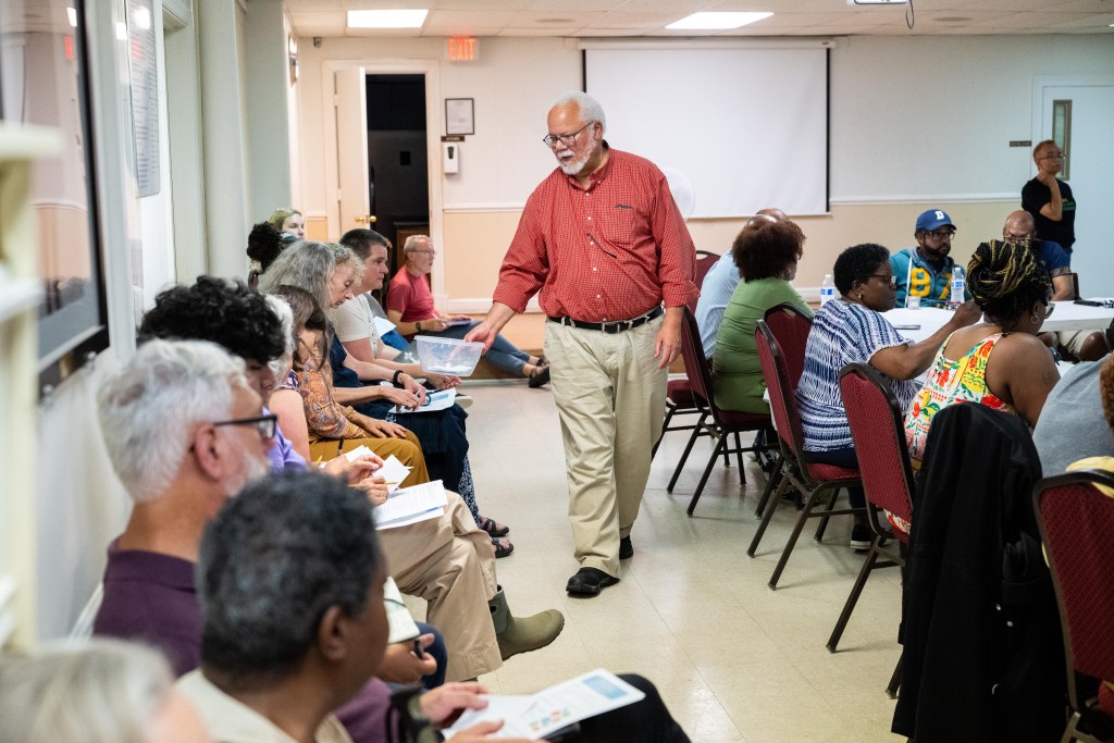 A photograph of a community meeting in a well-lit church basement. To the left of the photo, about a dozen people sit in chairs along a wall covered in framed photos and documents. To the right of the photo, about half a dozen people are seated at tables. A Black man with grey hair, a beard and glasses walks down the aisle between them, a plastic tub in his hand. There are a few index cards in the tub.