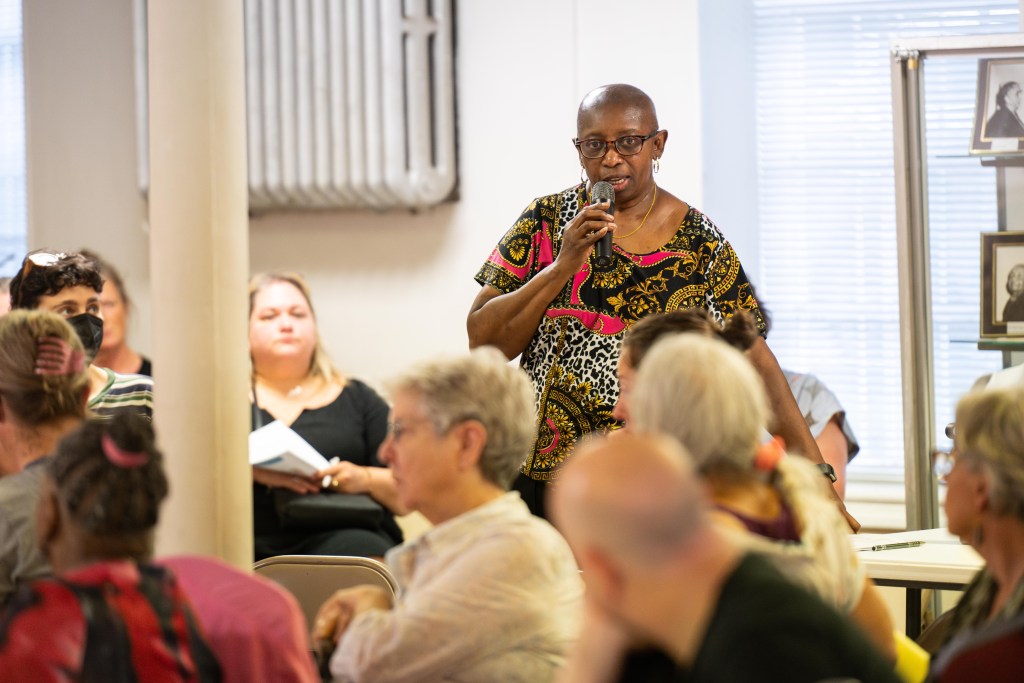 A photograph of a few people gathered at a meeting in a church basement. The photograph focuses on an older Black woman wearing glasses, hoop earrings and a colorful patterned top. She is holding a microphone and speaking to the crowd.