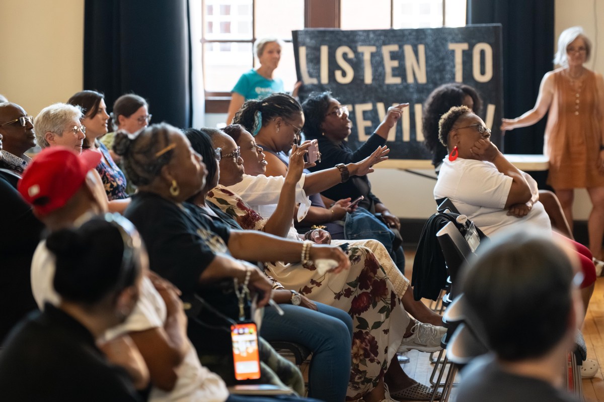A photograph of a multi-racial group of adults, most of them Black women, sitting in plastic chairs in an auditorium, facing a stage that is out of the frame. Some of the women in the photo are talking and pointing their fingers at people not visible in the photo. In the background two white women hold up a banner that says "Listen to Fifeville!"