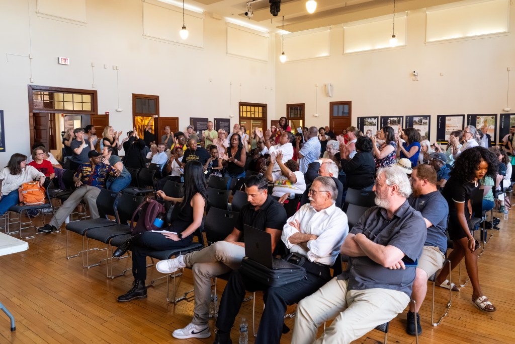 A photograph of a multi-racial group of more than 100 people at a community meeting in a brightly-lit auditorium. Most of the people in the photo are standing and clapping. A few people, seated in the front, sit with their arms across their chests.