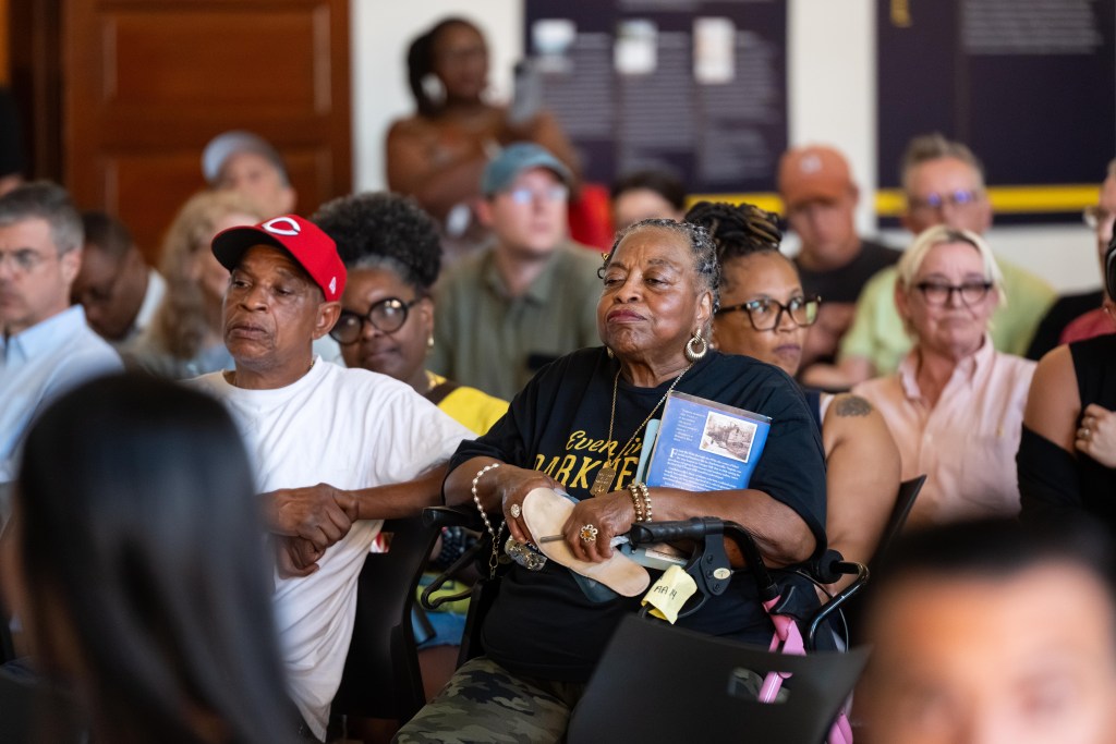 A photograph of people gathered in an auditorium. At the center of the photo are two older Black people, a man and a woman. The man is leaning back in his chair, his eyes cast down to the floor. The woman is wearing a t-shirt that says "Even in the darkness I see His light" and is sitting in a rollator. Her arms are in her lap and her lips are pursed in disapproval.