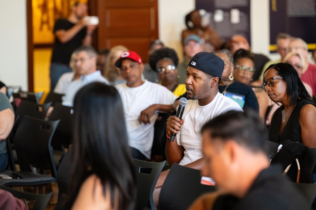 A photograph of a community meeting in an auditorium. At the center of the photo is a Black man wearing a backwards baseball cap and speaking into a microphone.