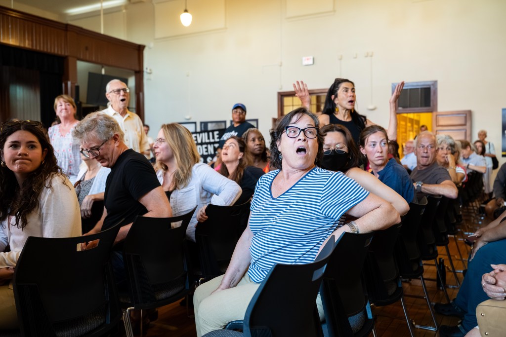 A photograph of about two dozen people at a community meeting in an auditorium. Most of the people are white, though some are Black. All are reacting to something someone out of the frame is saying. Multiple people are grimacing while others' mouths are gaping open. One woman is standing up with her mouth open and her arms outstretched. One woman rolls her eyes.