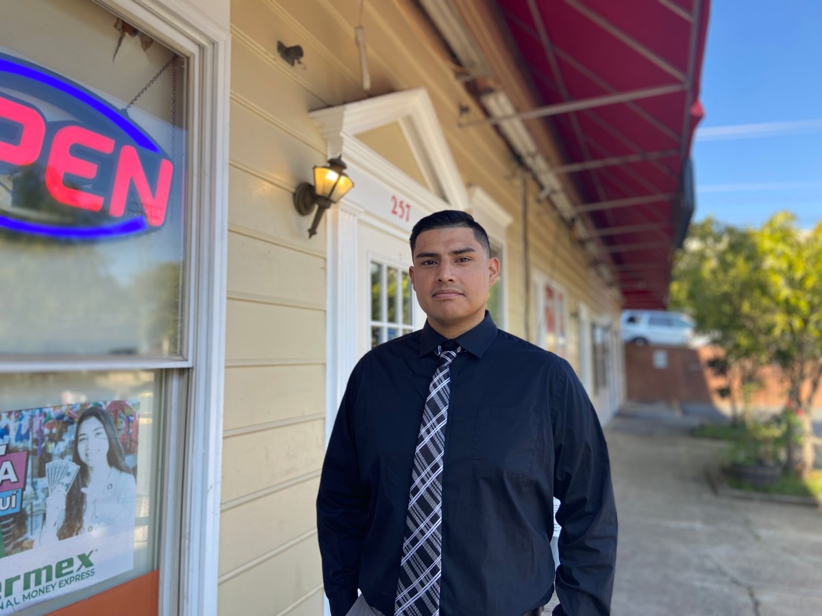 A man in a black shirt and tie stands in front of a store with part of a neon "Open" sign in the foreground.