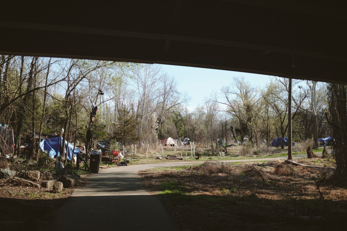 A view from under a bridge, with a wooded trail winding into the distance. The bridge looms overhead and casts a large shadow across the foreground. Beyond the trail are several tents and other items from people camping at the site.