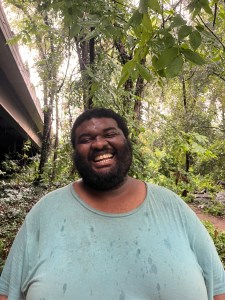 A man in a light green shirt smiles, foliage and the side of a bridge in the background behind him.