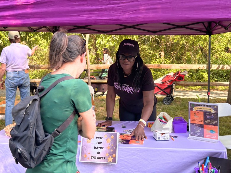 A woman stands under a purple canopy behind a table with a purple tablecloth. She leans in to talk with a woman who has come up to her table.