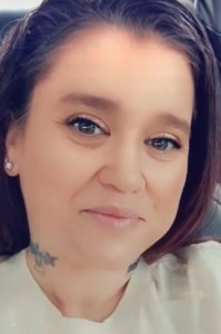 A close up shot of a woman smiling at the camera. She is wearing white earrings and a cream t-shirt, with a tattoo on her neck.