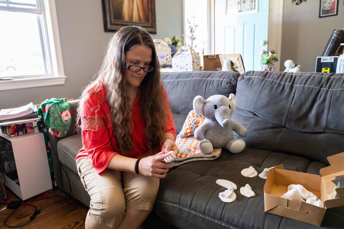 A woman with long hair and glasses sits on a couch in her home, surrounded by a stuffed elephant, a colorful blanket, and plaster casts of a baby's hands and feet. She holds a cast of a baby's hand and rests her index finger on the palm of the tiny hand.
