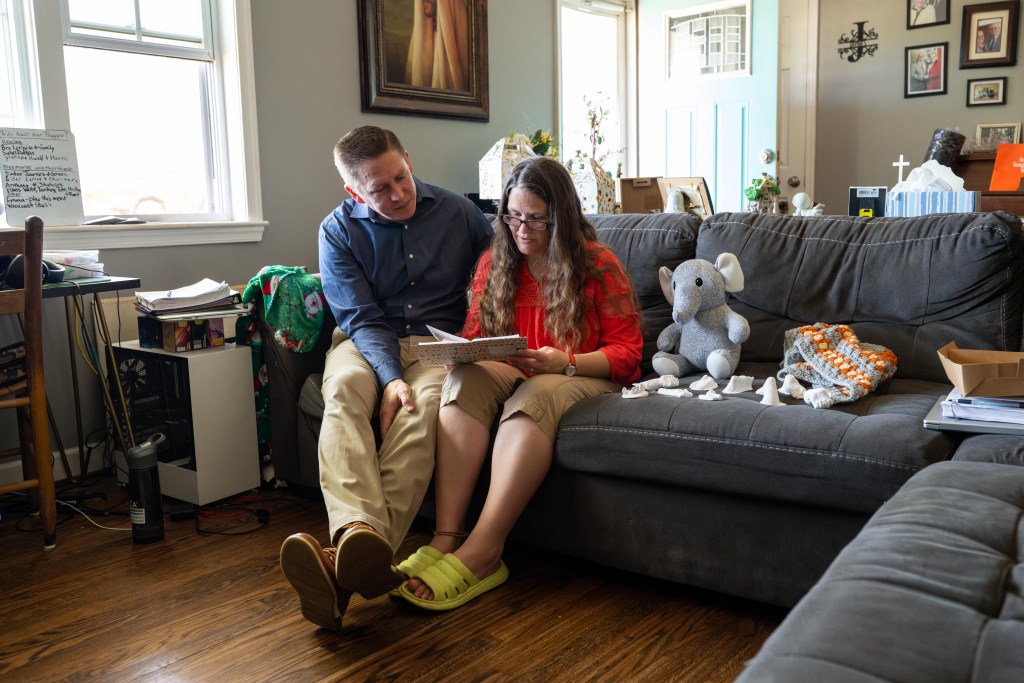 A man and woman sit together on a couch, looking at a photo album, with a stuffed elephant, small plaster hand and foot casts, and a crocheted blanket beside them.