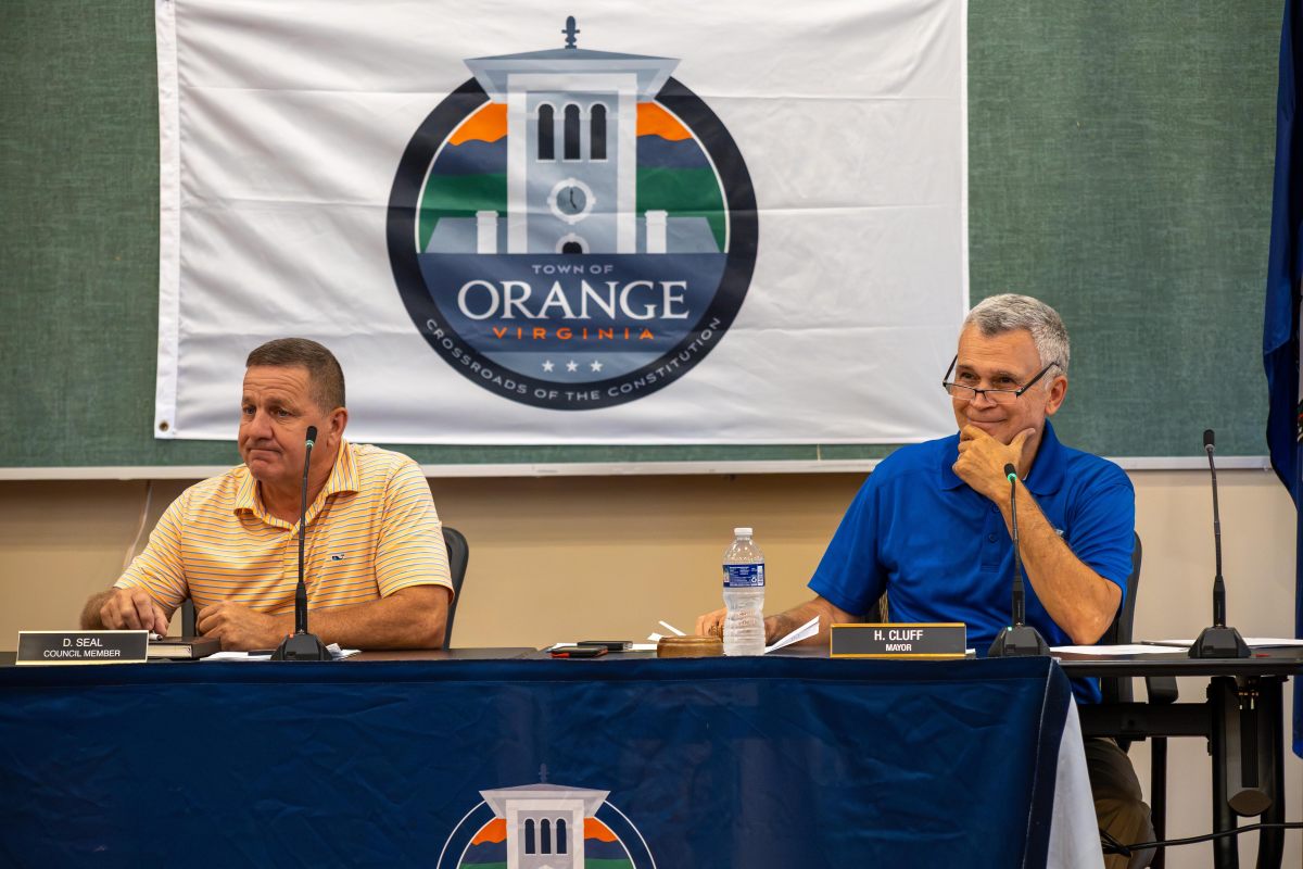 Two men sit at a conference table with microphones and a dark blue table skirt. The man on the left is wearing a striped yellow polo and has a desk nameplate in front of him that reads "D. Seal Council Member." The man on the right is wearing a bright blue polo and the nameplate in front of him reads "H. Cluff Mayor." A White flag with a logo of the town of Orange is hanging on the wall behind them.