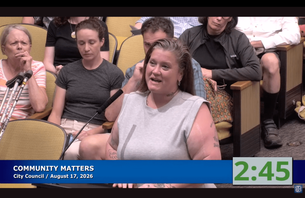 A woman at a microphone with people sitting in yellow chairs behind her. There is a chyron in a blue bar at the bottom that says "COMMUNITY MATTERS City Council / August 17, 2026" with a time stamp on the right that shows 2:45.