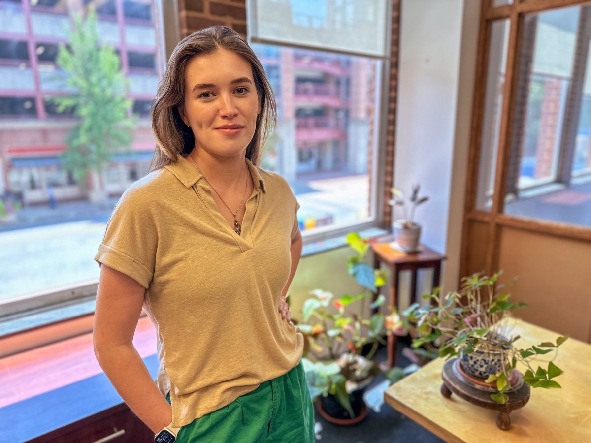 A woman smiles at the camera. She stands in an office with large windows and plants.