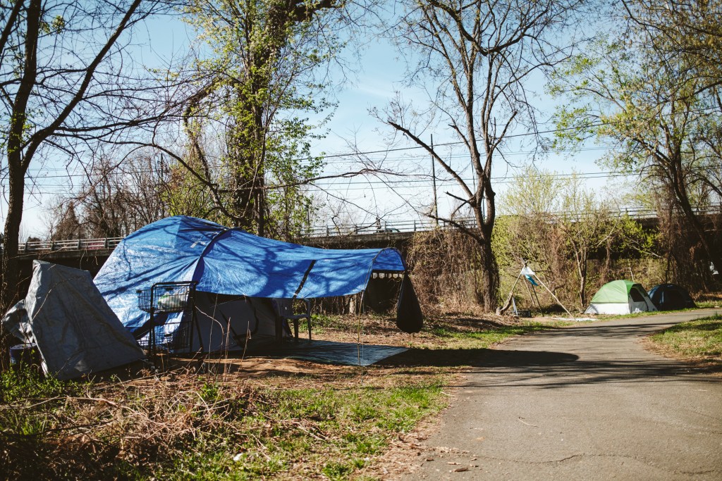three tents in a homeless encampment along a paved public trail. The trees are starting to bud, and the ground around the trail is a mixture of green and brown. There is a bridge in the background