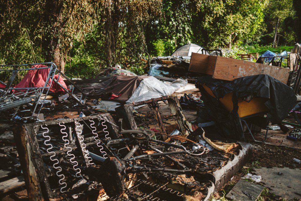 The aftermath of a fire, with charred bed frames, a burned bicycle and burned debris lying scattered across the ground in a wooded area.