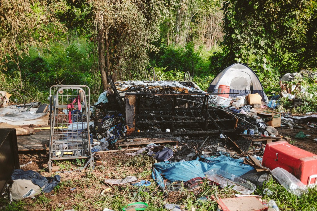 Burned furniture frames, a large cooler and scattered supply boxes sit among debris near trees.