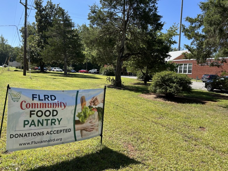 A banner reading "FLRD Community Food Pantry, Donations Accepted" stands on a lawn in front of a brick school administration building.