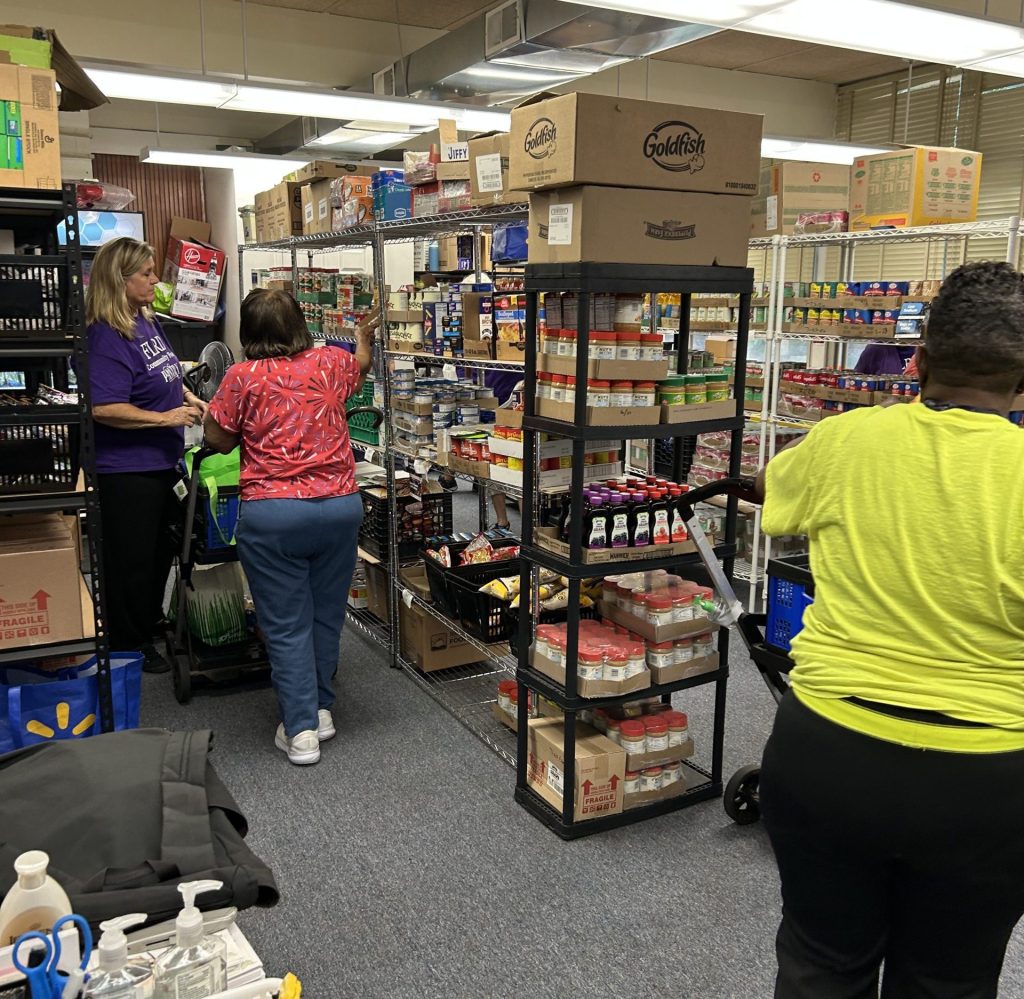 Volunteers and shoppers browse shelves stocked with canned goods and packaged food inside a food pantry storage room.