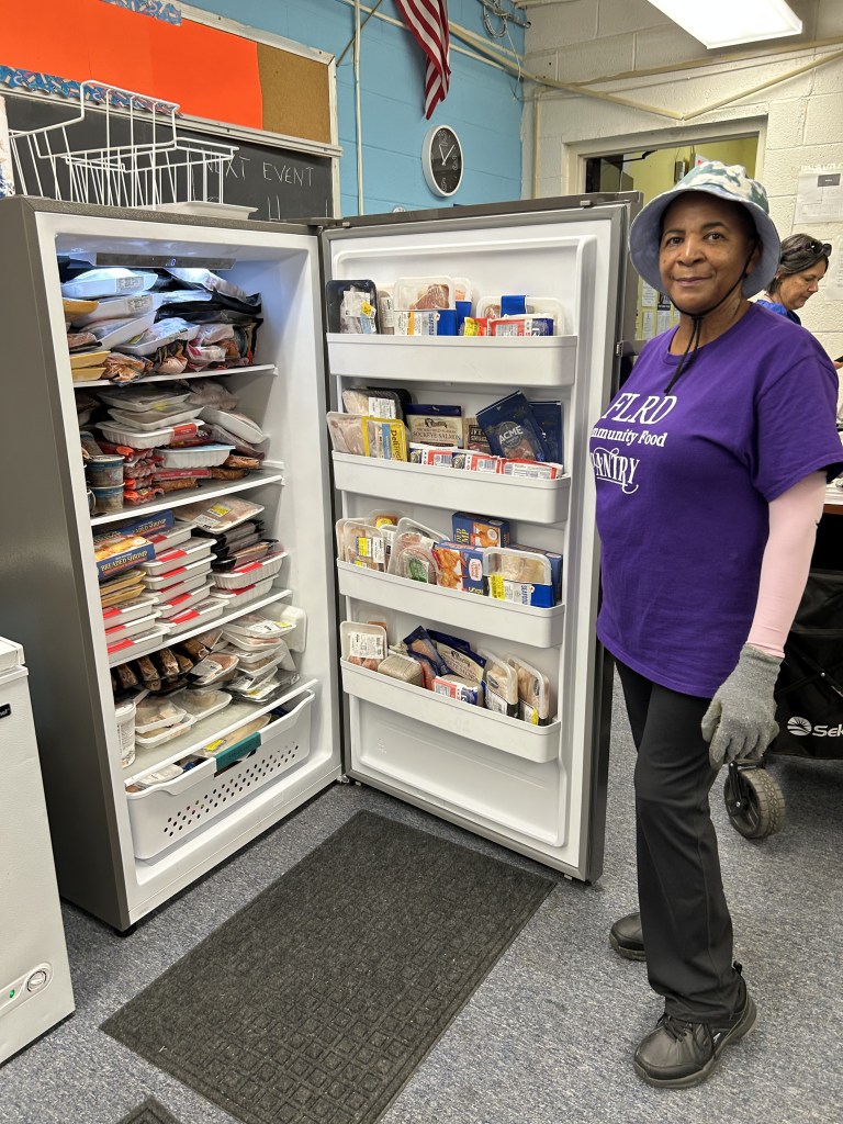 A woman wearing an "FLRD Community Food Pantry" shirt stands beside an open freezer stocked with packaged meat and frozen foods.