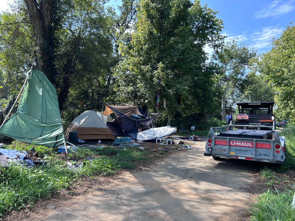 A walking trail runs through a wooded area, with three abandoned tents visible on the left-hand side next to the trail. A small utility vehicle with a trailer sits on the right-hand side of the trail. Several black footlockers are loaded onto the trailer.