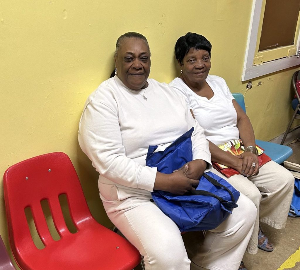 Two women sit together in chairs against a yellow wall, one holding a blue tote bag.