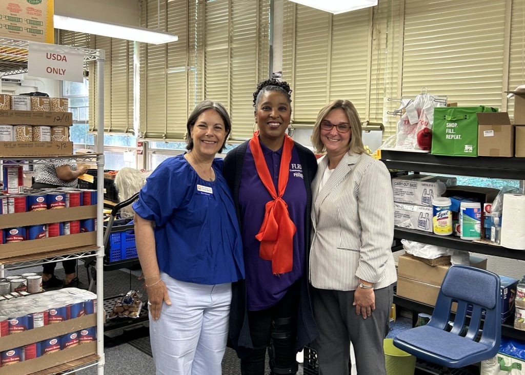Three women stand smiling together in a food pantry storage room lined with shelves of canned goods.