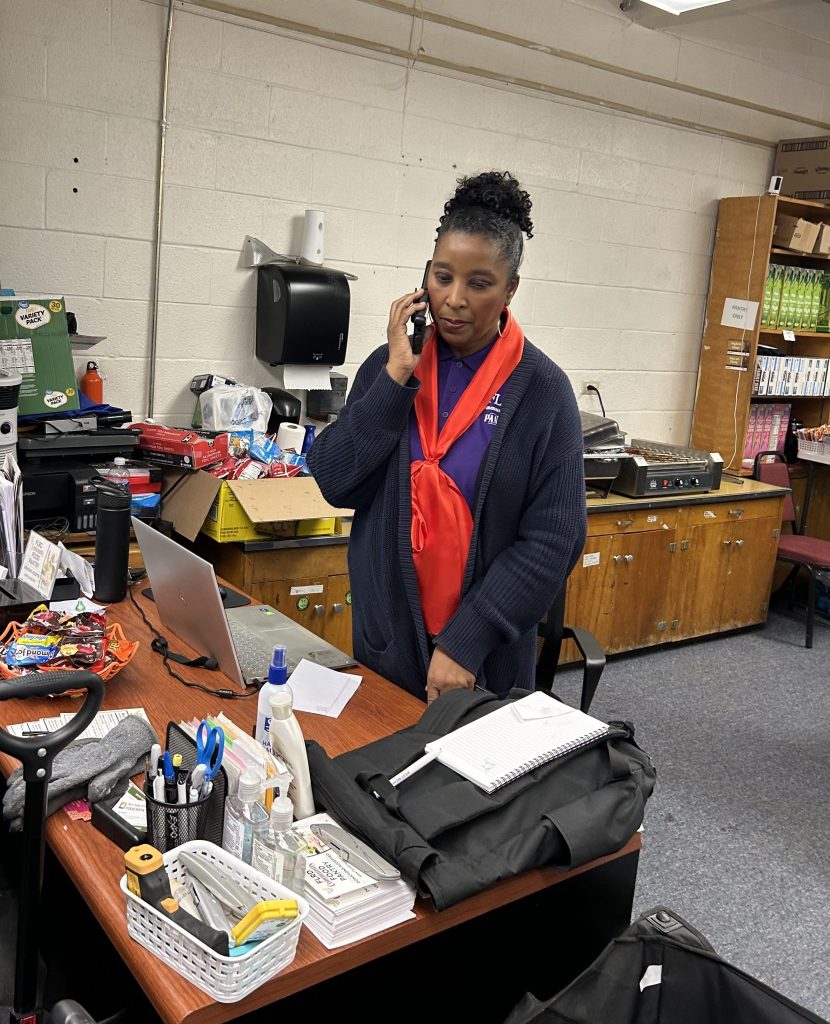 A woman talks on a phone while standing at a desk with a laptop, supplies, and a rolling cart in a pantry office.