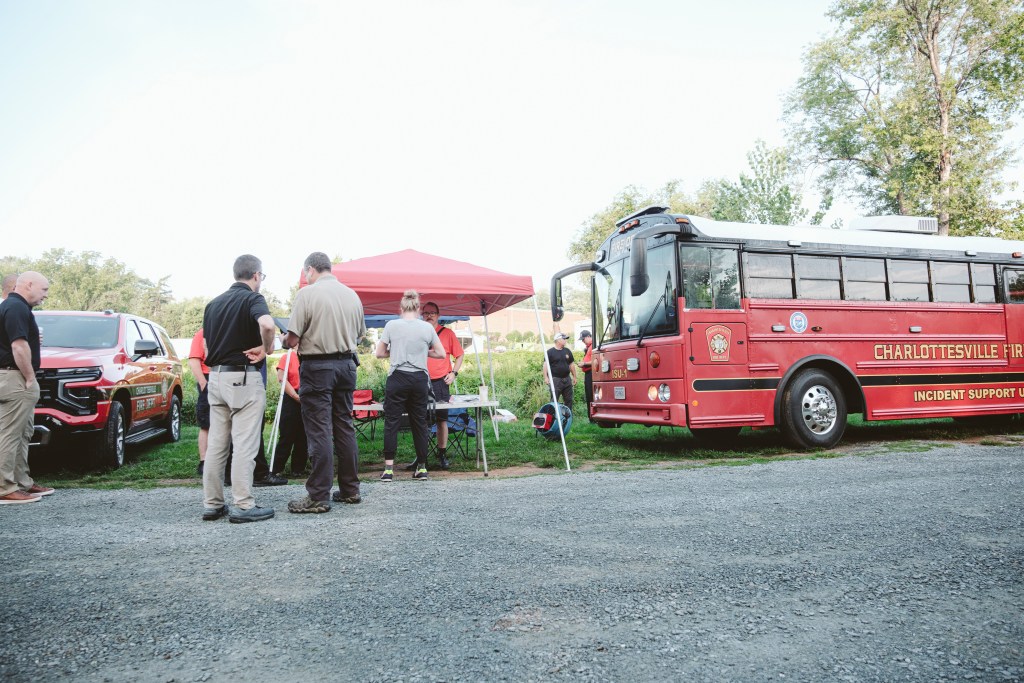 A red Charlottesville Fire Department vehicle and support bus are parked near a canopy tent where several people stand talking.