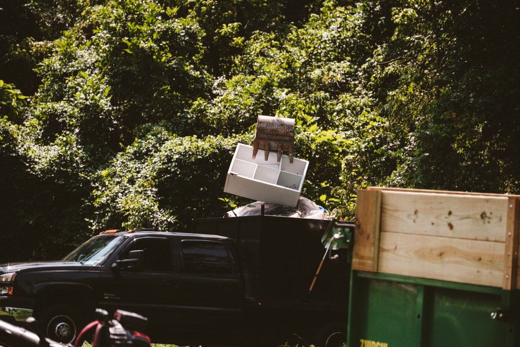 A wooded trail, flush with late-summer vegetation. An excavator picks up a six-cube shelving unit in its bucket, preparing to dump the piece of furniture into the bed of a truck, on top of a tent. In the front right corner of the photo there is a large metal dumpster with some sort of wood furniture on top.