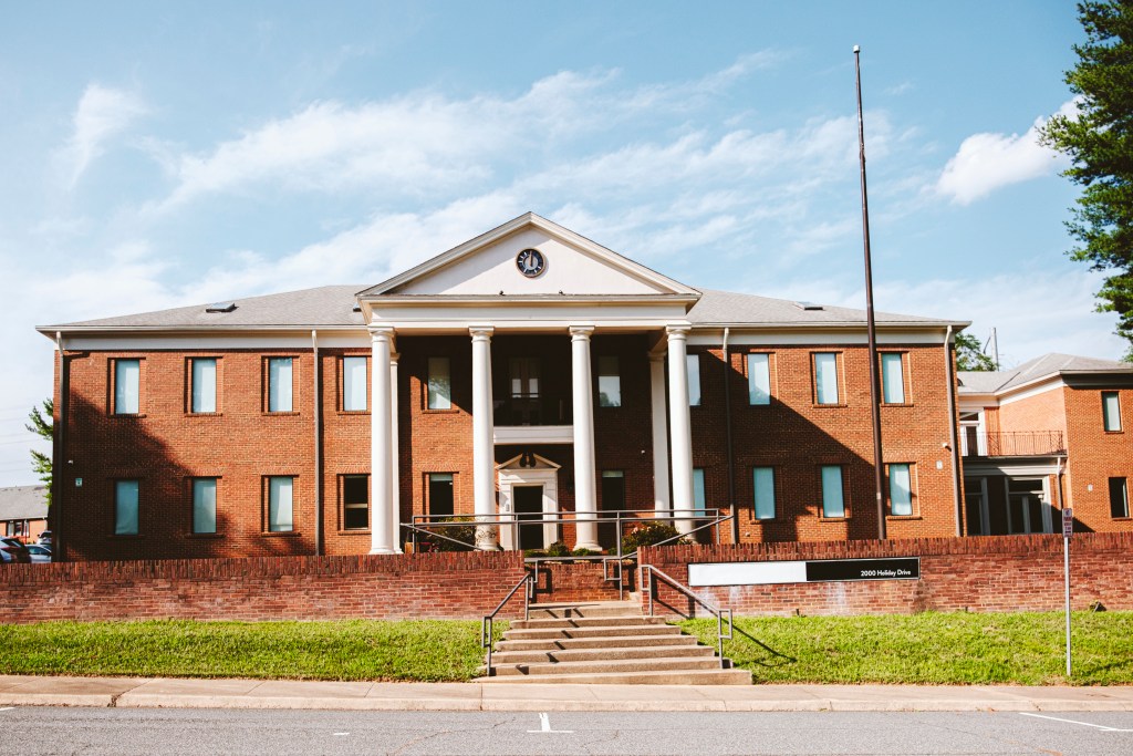 A two-story brick building with white columns near the front entrance. Concrete stairs lead up to a brick wall, which encloses a patio area in front of the building.
