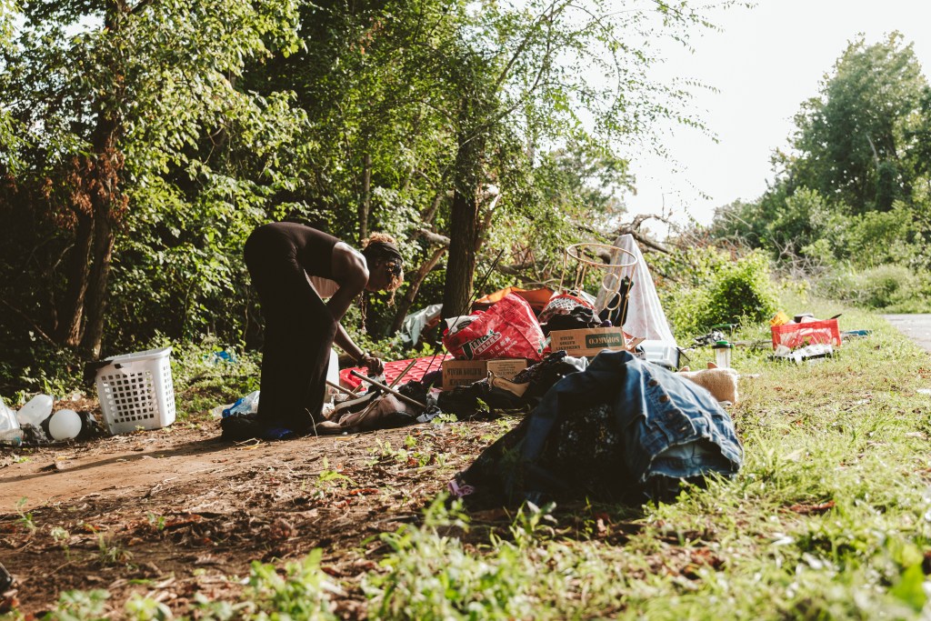 A woman in a dress bends over, sorting through scattered belongings, including boxes, bags, and a laundry basket, on a wooded path.