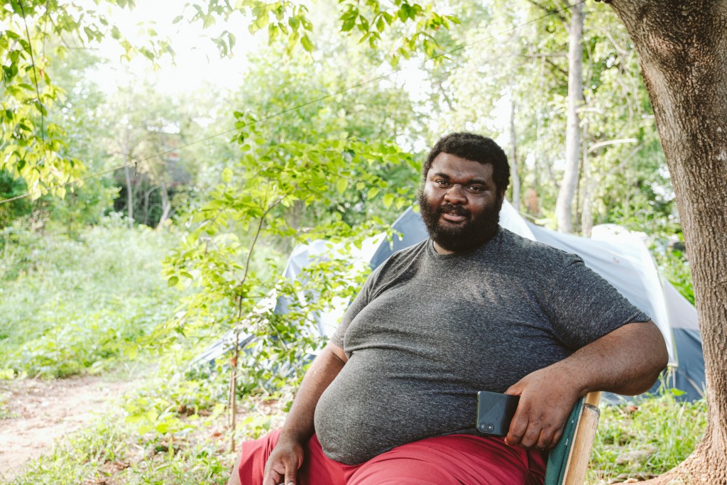 A man sits in a chair. His left arm is propped up on the back of the chair, and he holds a cell phone in his left hand. He wears a t-shirt and pants rolled up to his knees. Behind him is a three-domed tent and abundant green foliage.