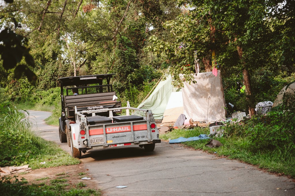 A utility vehicle towing a "U-Haul" trailer drives along a paved path near tents and scattered belongings in a wooded area. Black footlockers are visible in the trailer bed.
