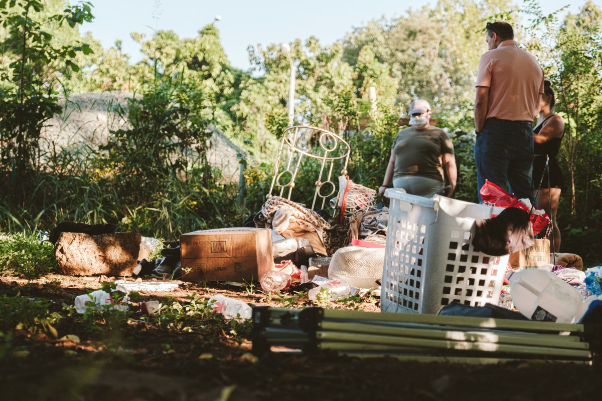 A photograph of a person's belongings scattered around on the ground at a homeless encampment along a public trail. Three people stand around, looking at the items, which include a metal-framed round side table, a cardboard box tipped over on its side, a plastic laundry basket, and various clothes, blankets, and empty plastic bottles.